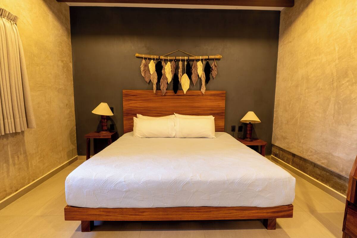 Una cama bien tendida con cabecera de madera, dos mesitas de noche con lámparas y un adorno en la pared sobre la cama.