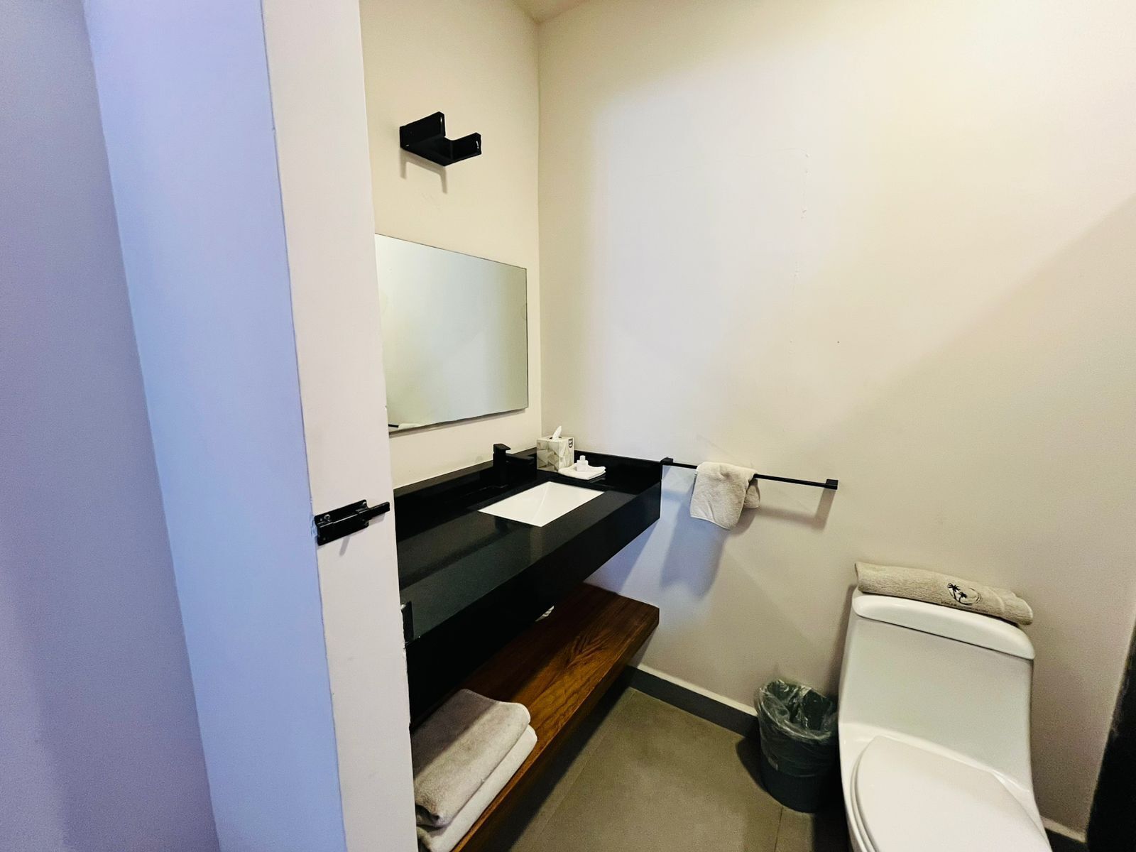 Uma casa de banho pequena com sanita, lavatório com espelho, porta-toalhas e um contentor de lixo.