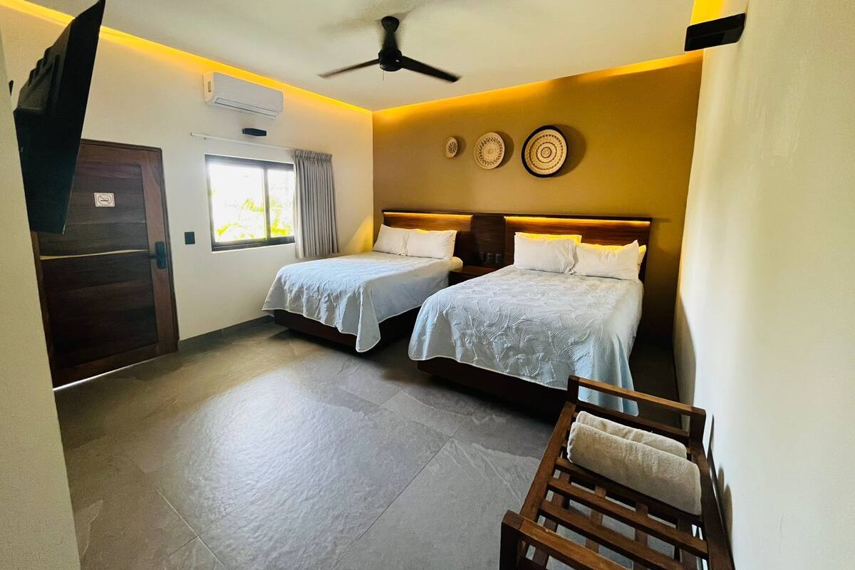Habitación de hotel con dos camas, un ventilador de techo, una ventana con cortinas, una puerta y un aire acondicionado instalado en la pared.