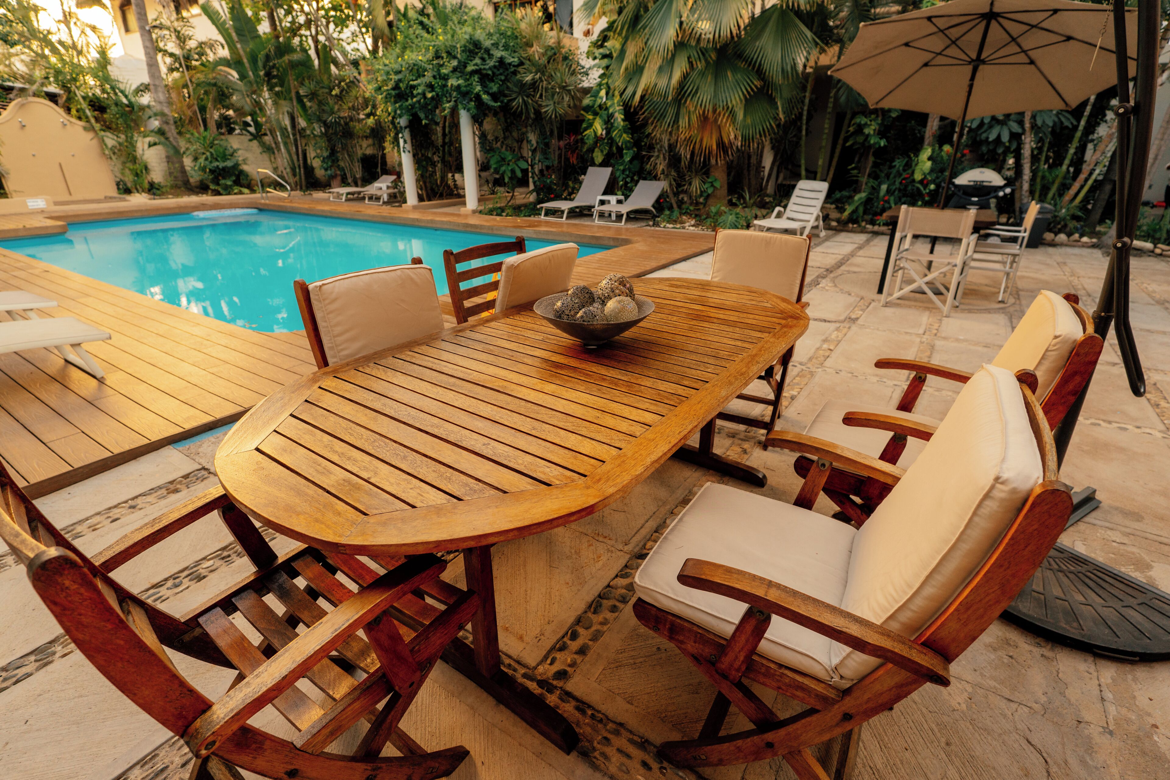 Une table en bois placée au bord de la piscine, avec des chaises, un saladier rempli de fruits et une piscine en arrière-plan.