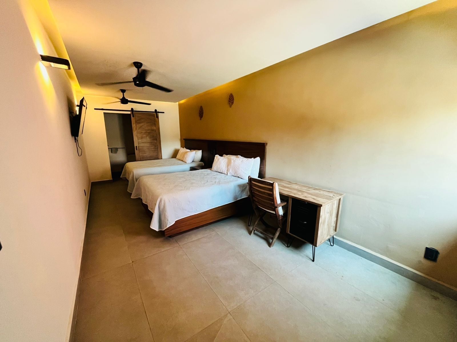 Ein Schlafzimmer mit zwei Betten, einem Schreibtisch, einem Stuhl, einem Deckenventilator und einem an der Wand befestigten Telefon.