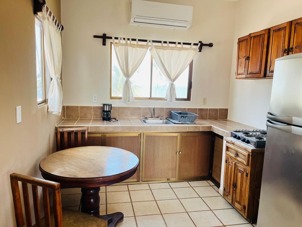 A compact kitchen with a round table, wooden cabinets, a built-in gas stove, and a window with curtains.