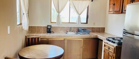 A compact kitchen with a round table, wooden cabinets, a built-in gas stove, and a window with curtains.