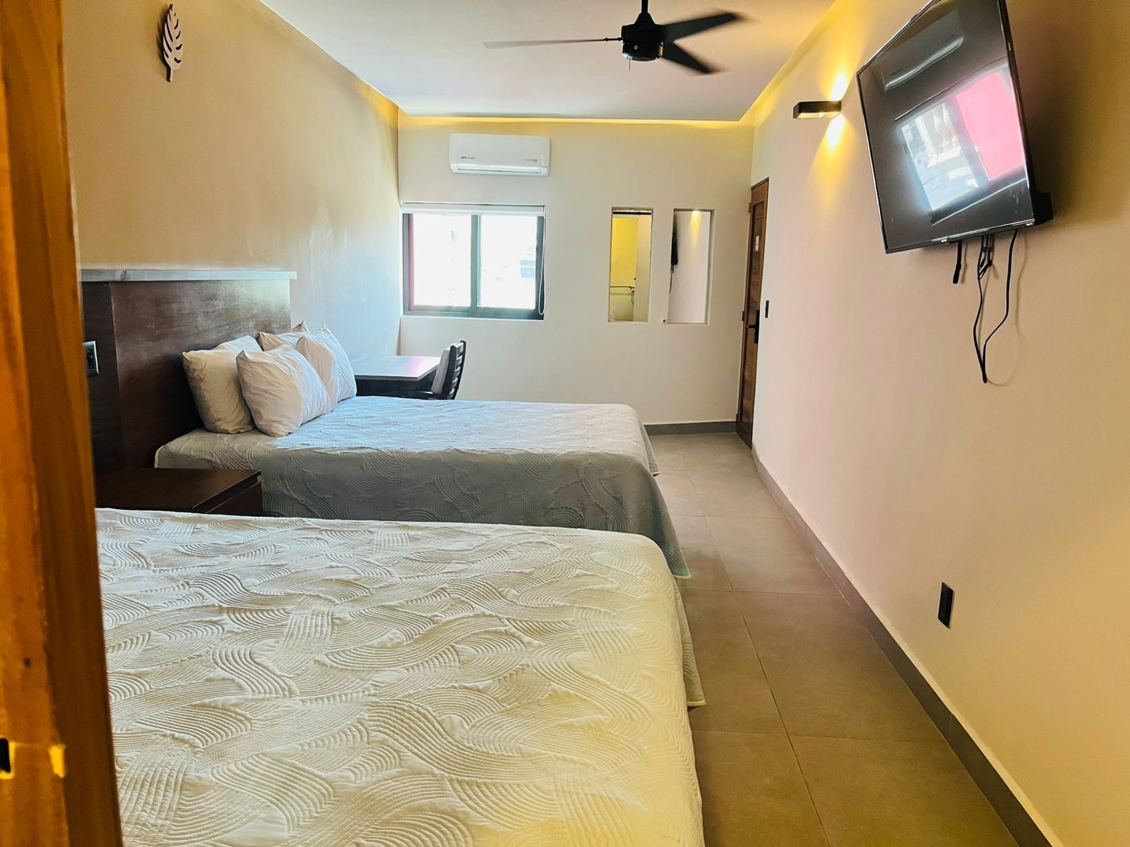 A hotel room with two beds, a ceiling fan, a TV, and an air conditioning unit.