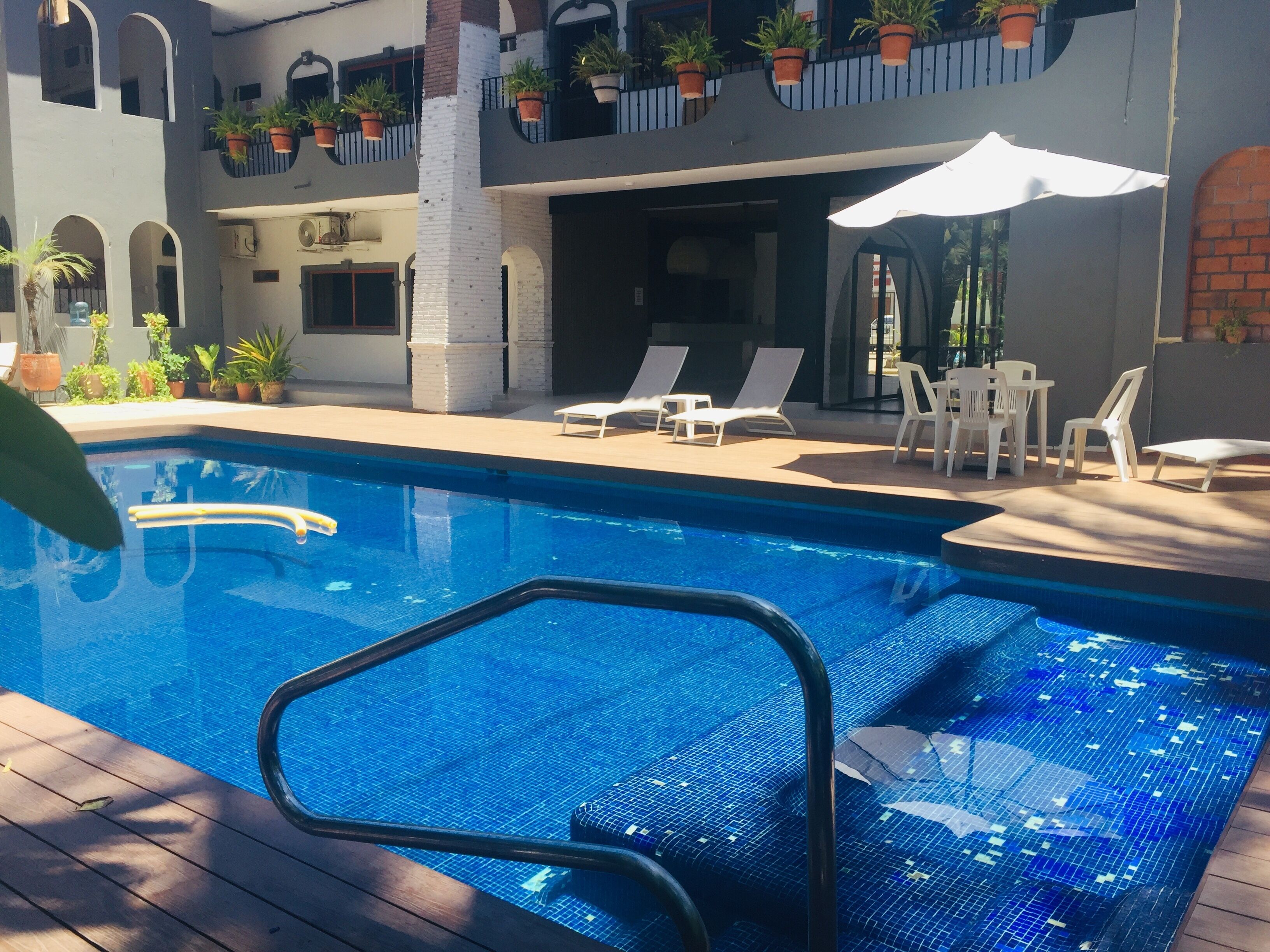 Zona de piscina con sillones de descanso y sombrillas, rodeada de plantas en macetas y un edificio con balcones.
