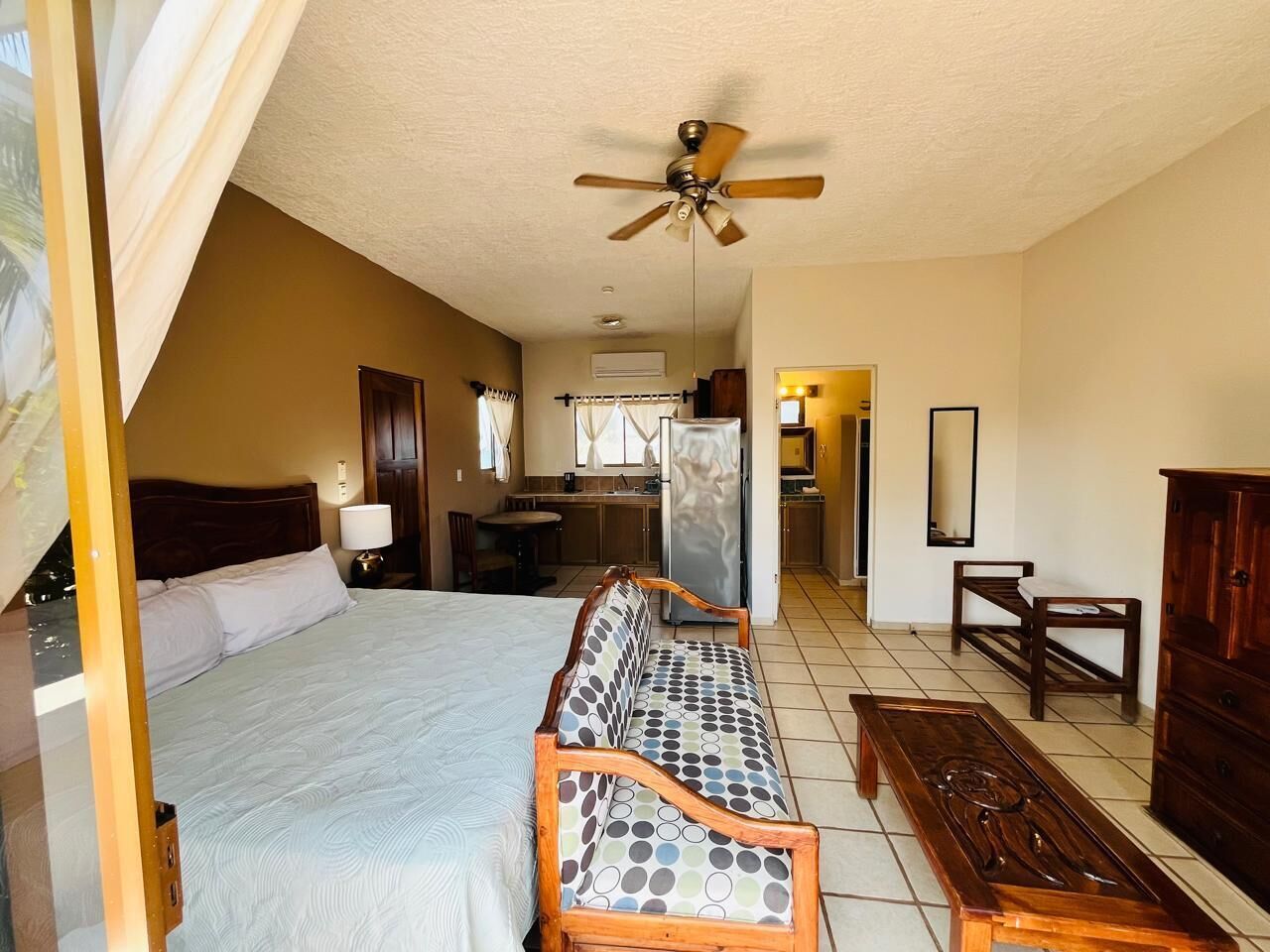 A bedroom with a bed, a sofa, a wooden bench, a ceiling fan, and a kitchenette area.
