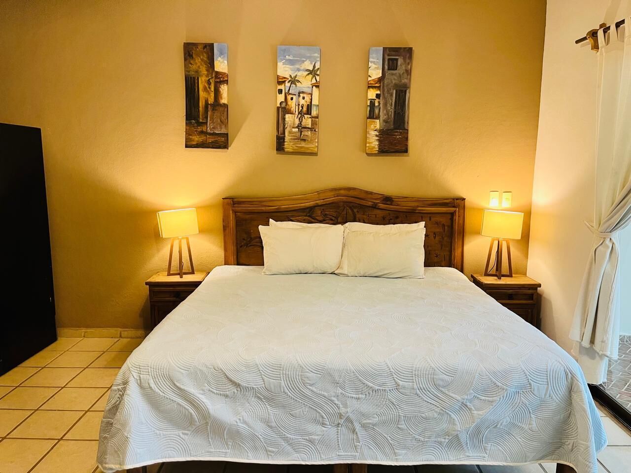 Habitación de hotel con una cama grande, dos lámparas de noche y tres cuadros en la pared.