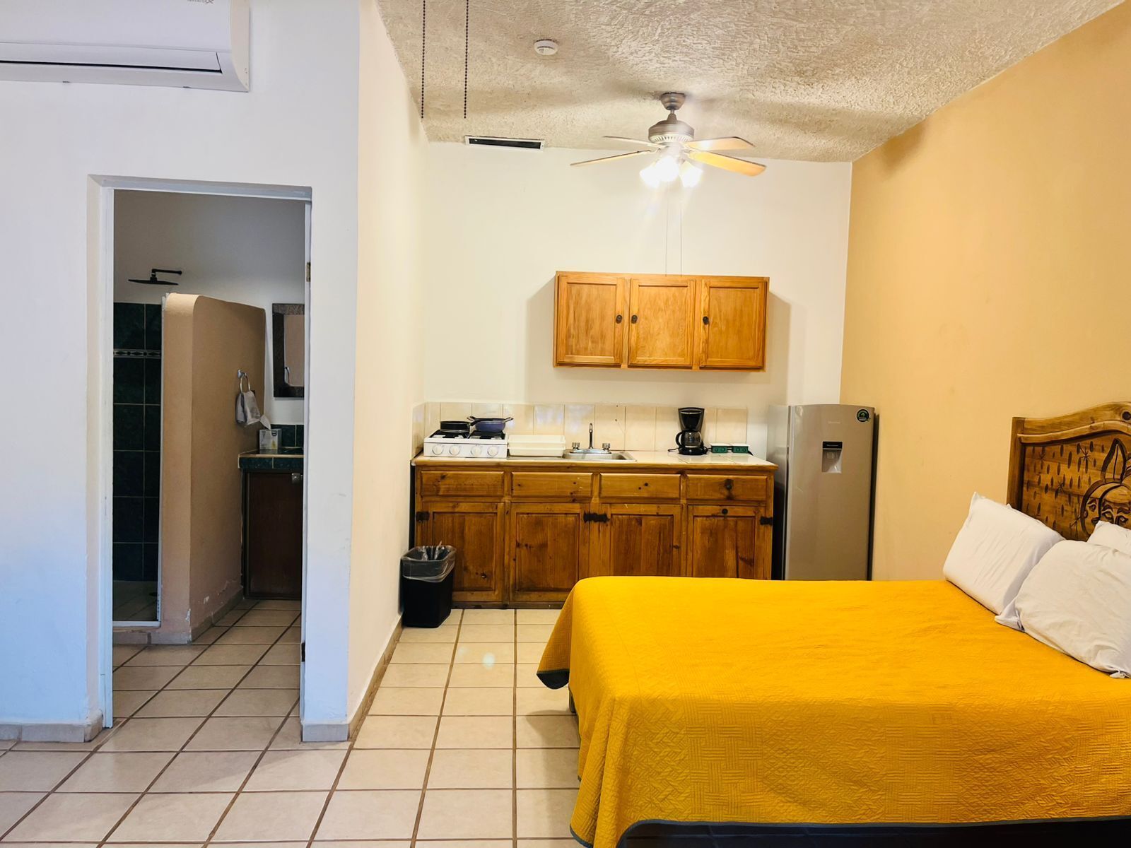 Een kleine kamer met een bed, een kitchenette, een plafondventilator en een deur naar een badkamer.