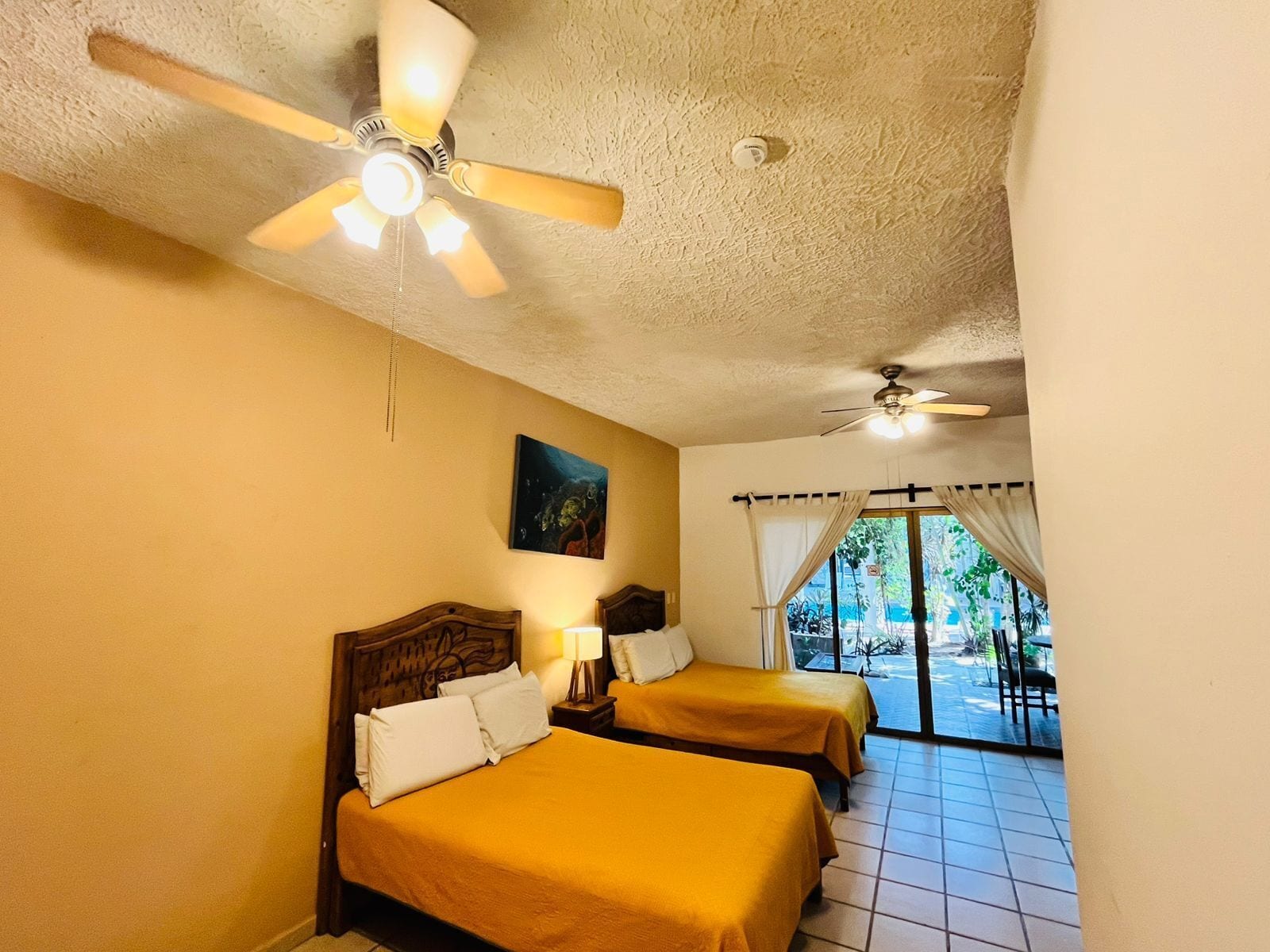 Habitación de hotel con dos camas, ventilador de techo, un cuadro y vistas al exterior.