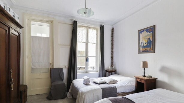 A hotel room with two beds, a wardrobe, a chair, a window with curtains, a lamp, and a poster on the wall.
