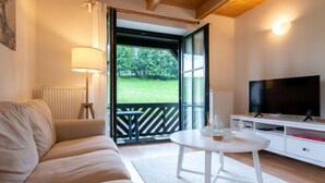 Un salon moderne avec un canapé, un téléviseur, une petite table et une vue sur un espace extérieur verdoyant, visible à travers une porte vitrée.