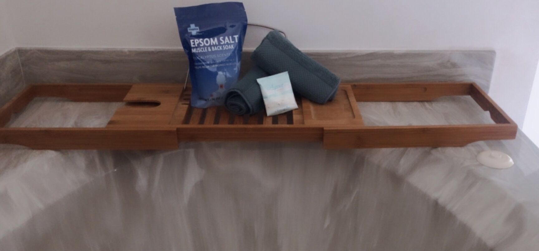 Un plateau en bois contenant un sachet de sel d’Epsom, une serviette roulée et un petit objet posé sur un lit.