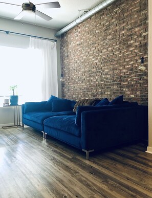 Relax in this cozy living space with a sofa against a brick wall, ceiling fan above, and a window with white curtains.