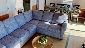 Un salon avec un canapé d’angle bleu, une table basse en bois agrémentée d’une plante, un coin repas avec une table et des chaises, et une cuisine visible à travers une porte ouverte.