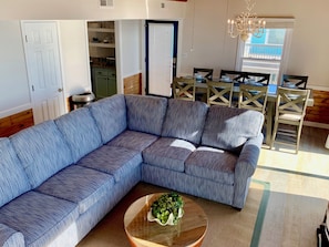 A living room with a blue sectional sofa, a wooden coffee table with a plant, a dining area with a table and chairs, and a kitchen visible through an open door. - Upscale Oceanfront, with Panoramic Views (Emerald Isle)