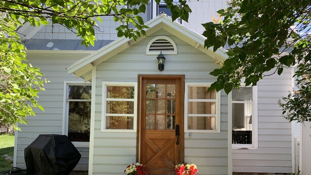 Une petite maison de plain-pied, dotée d’un porche à l’avant, d’une porte en bois et d’une petite fenêtre au-dessus de la porte. De chaque côté de la porte, on trouve deux plantes en pot.