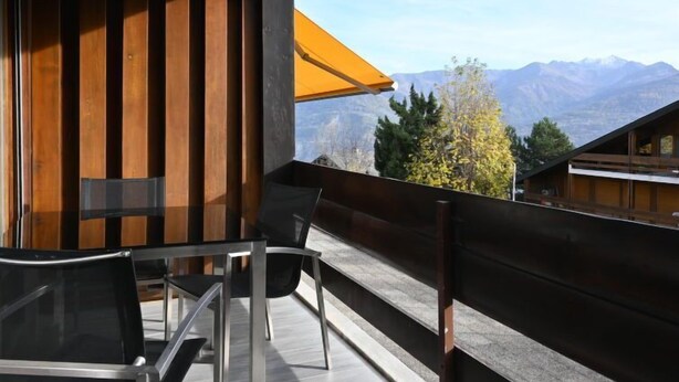 Ein Balkon mit Blick auf Berge, eine Holzwand und ein Glastisch mit Stühlen.