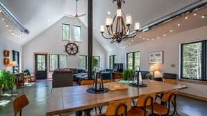 A spacious living area with a large wooden table, multiple chairs, a chandelier, and large windows offering views of the outdoors.
