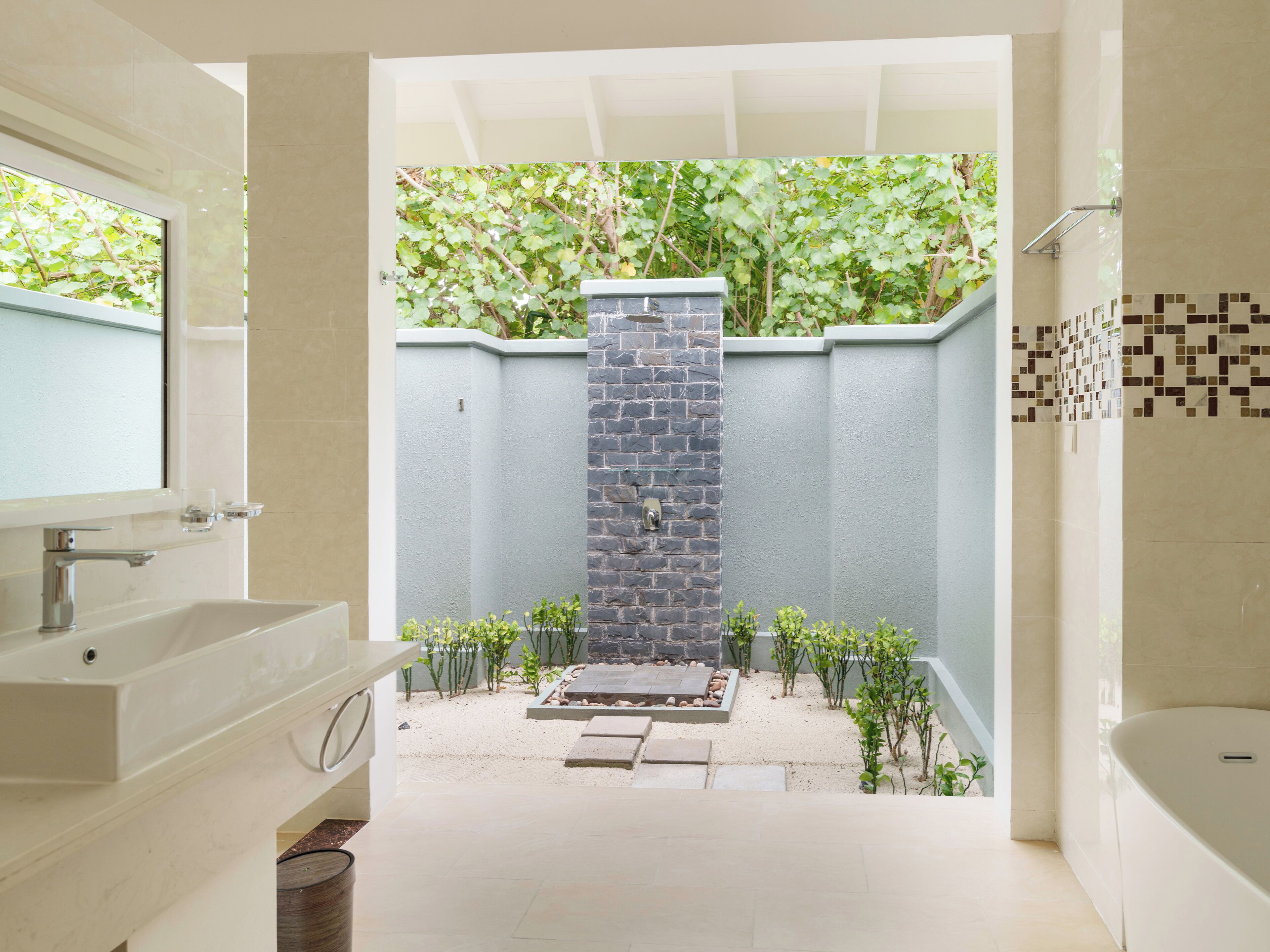 A bathroom with a large mirror, a sink, and a bathtub. There is a small garden area with plants and a stone wall in the background.