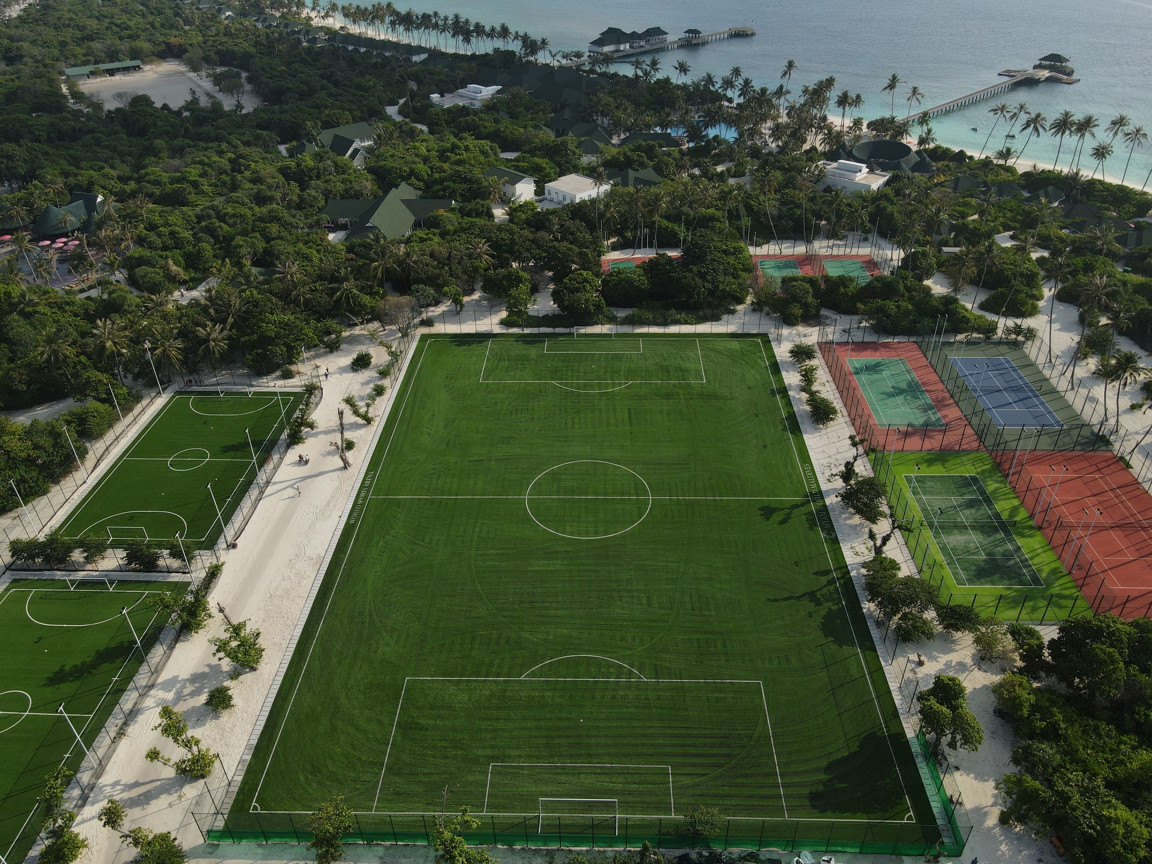 An aerial view of a sports complex with multiple courts, surrounded by lush greenery and a beach.