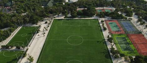An aerial view of a sports complex with multiple courts, surrounded by lush greenery and a beach.