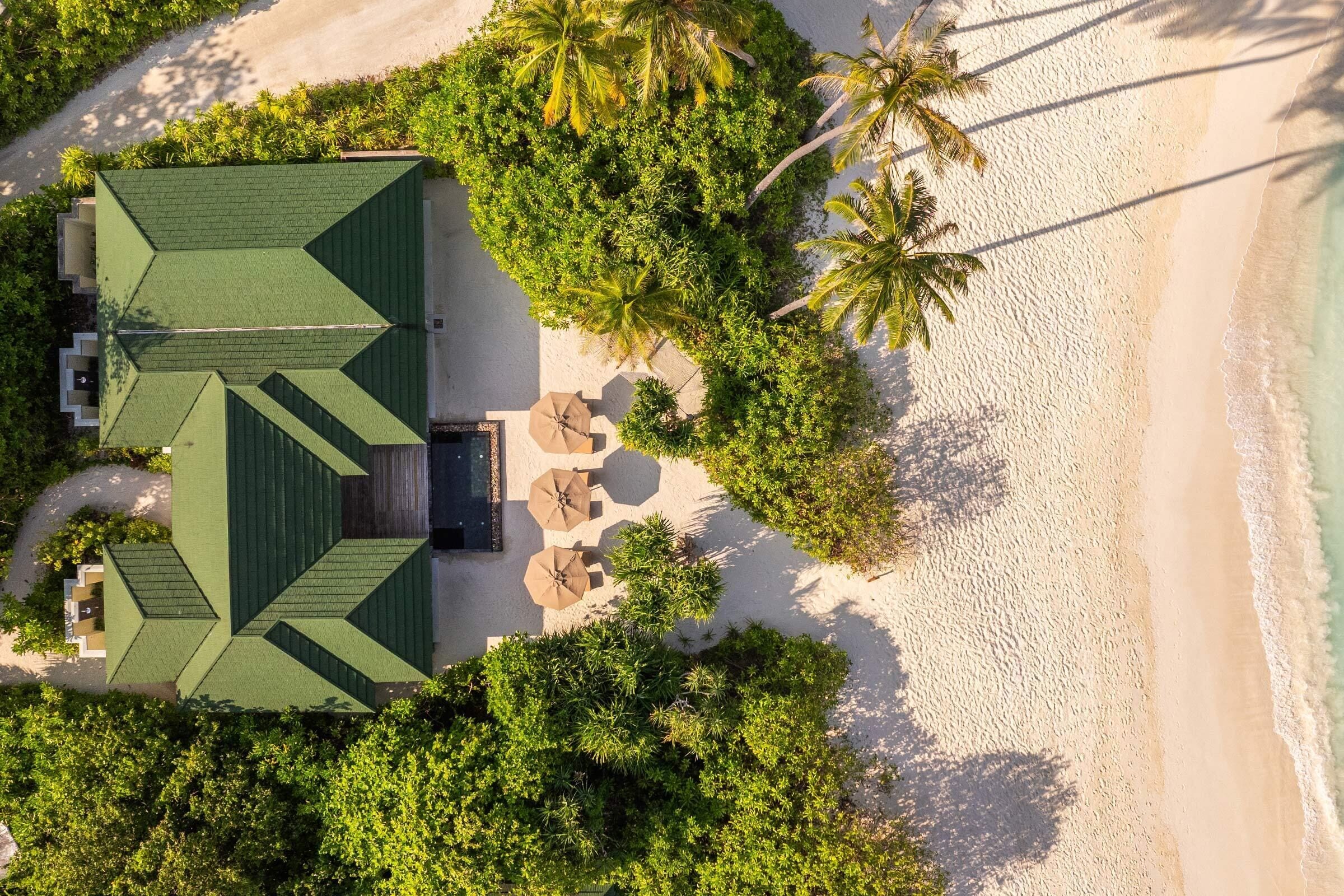 An aerial view of a resort with a green-roofed building, palm trees, and a sandy beach.