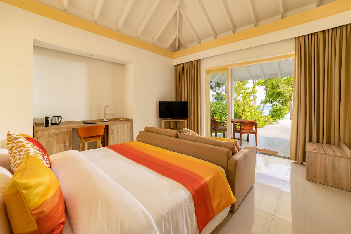 A hotel room with a bed, a sofa, a desk with a TV, and a view of greenery outside.