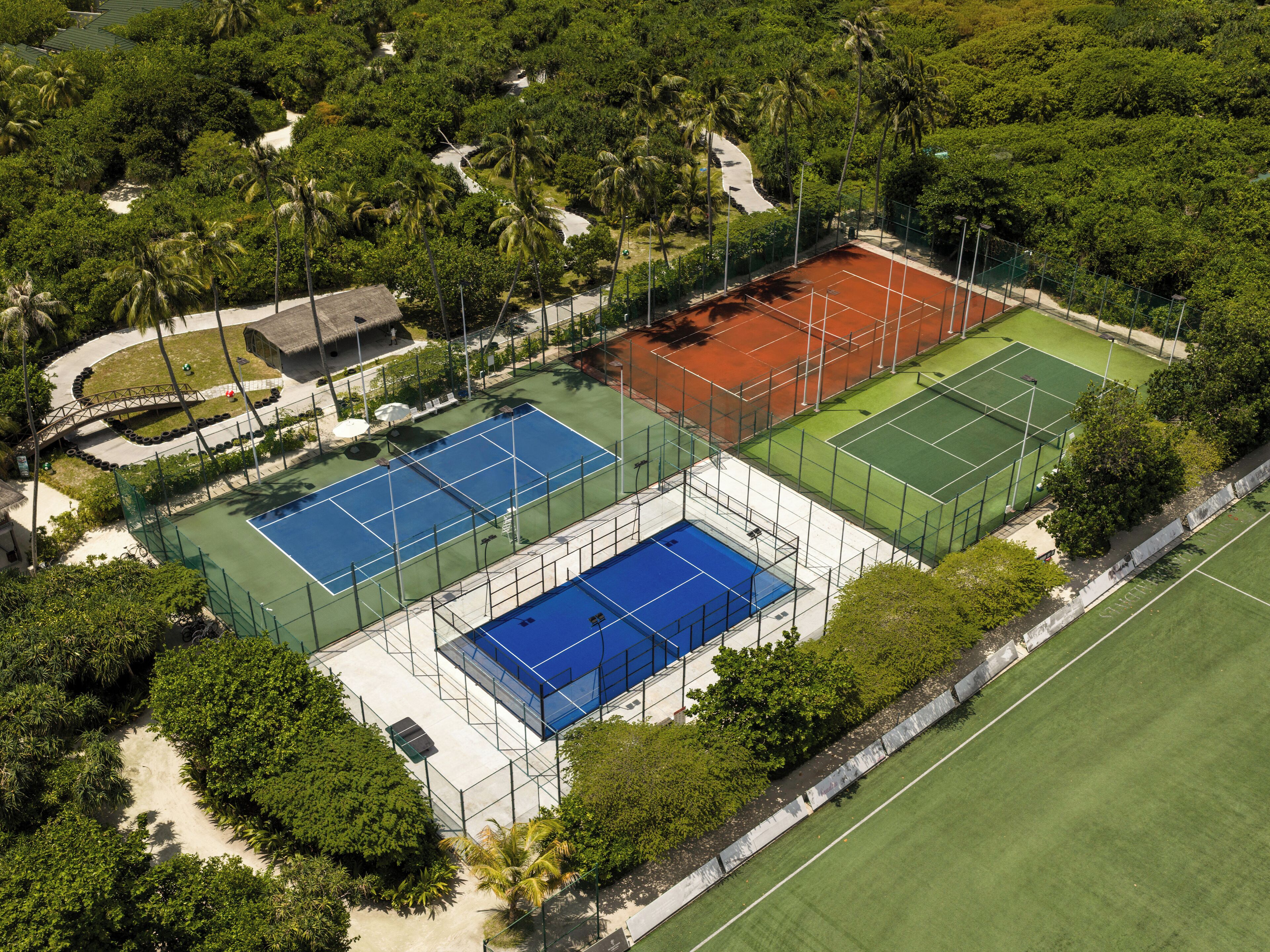 An aerial view of a tennis facility with multiple courts, surrounded by dense greenery and palm trees.