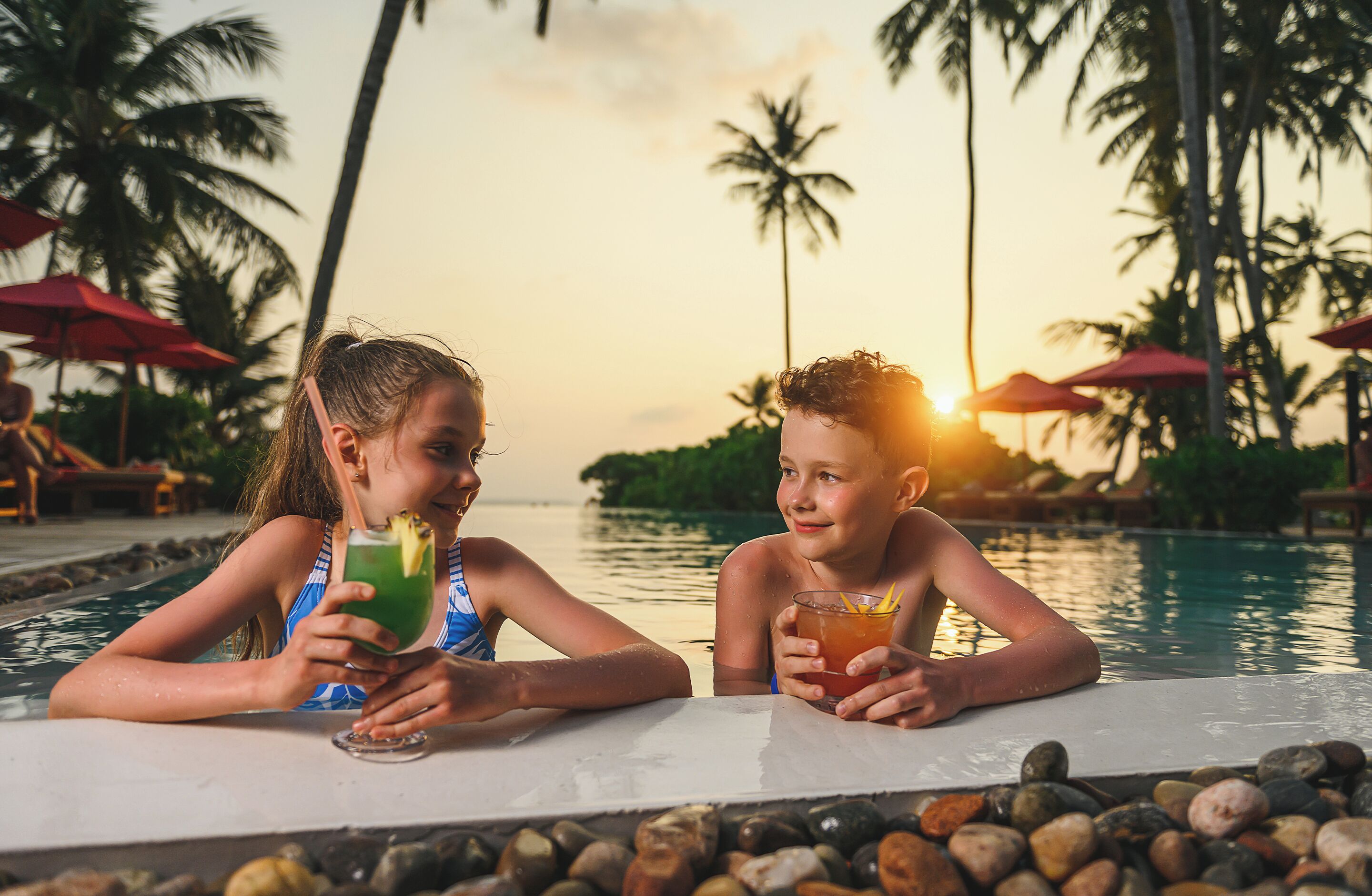Two people sitting by a pool with drinks, surrounded by palm trees and umbrellas.