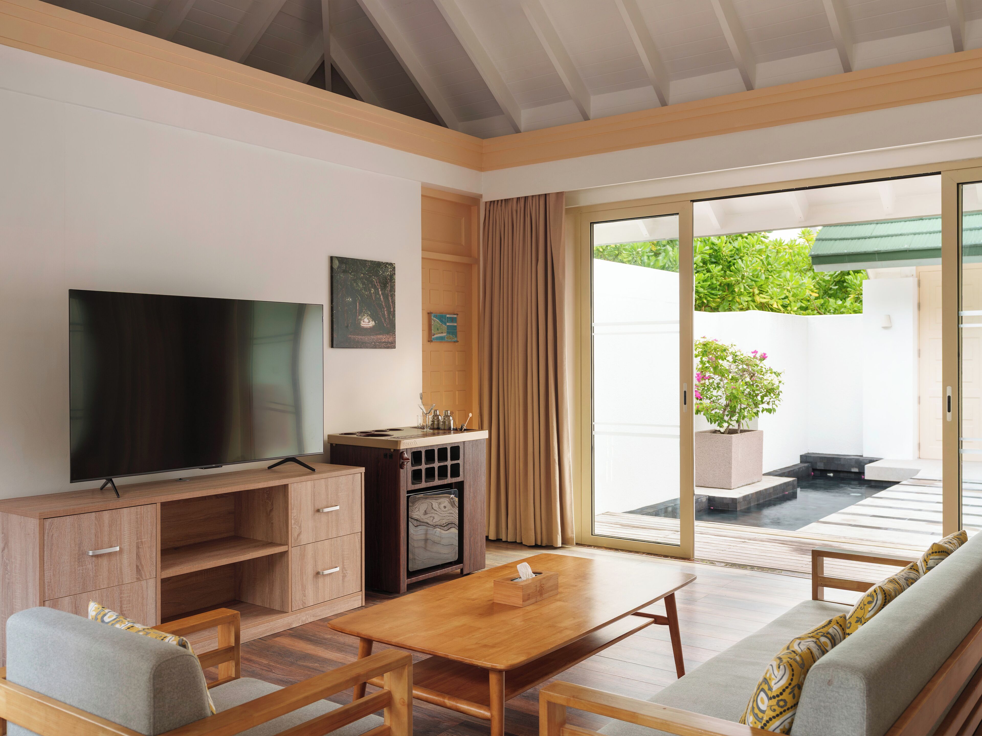 A modern living room with a flat-screen TV, wooden furniture, a coffee table, and a sliding glass door leading to a patio with a potted plant.