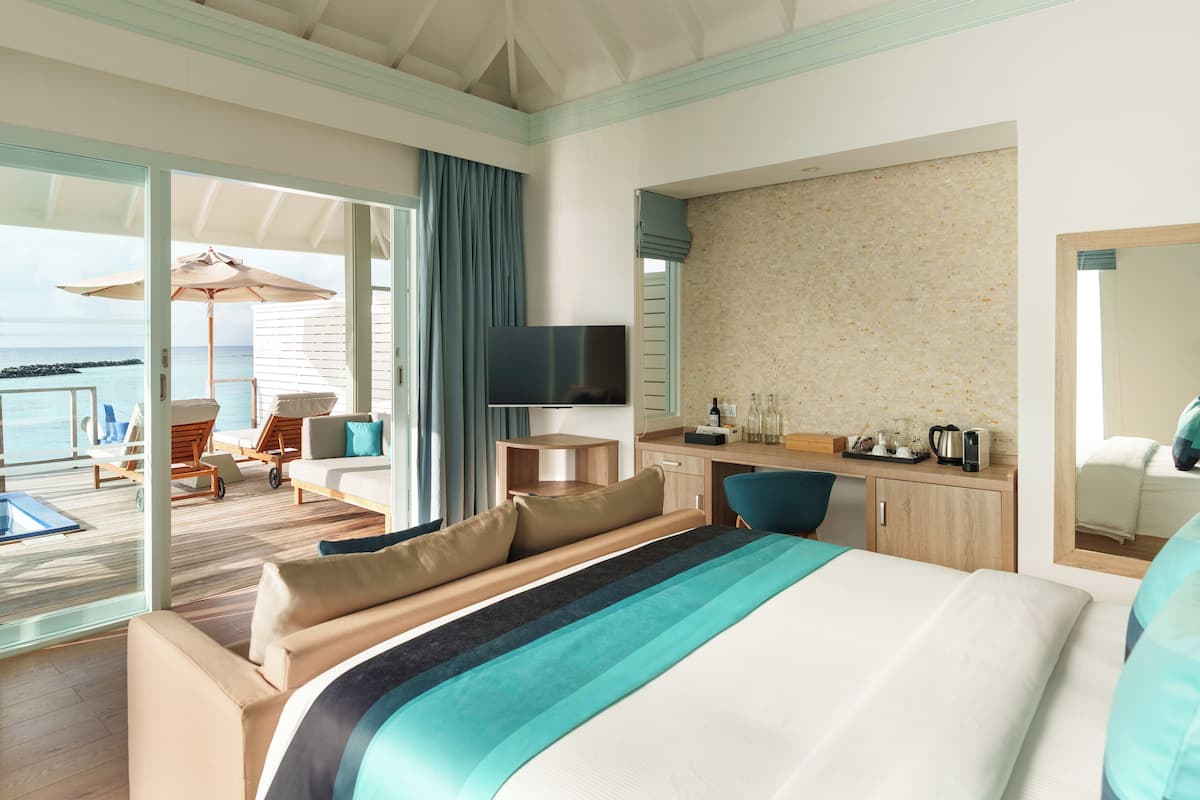 A hotel room with a large bed, a desk, a TV, and a balcony with a view of the sea.