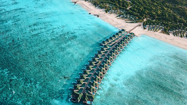 An aerial view of a resort with a long row of overwater bungalows connected by a walkway, surrounded by clear turquoise waters and a sandy beach. - Siyam World Maldives - All-inclusive  (Dhigurah)