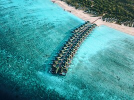 Vista aérea de un resort con una hilera larga de bungalows sobre el agua, unidos por un paseo marítimo, rodeado de aguas turquesas cristalinas y una playa de arena.