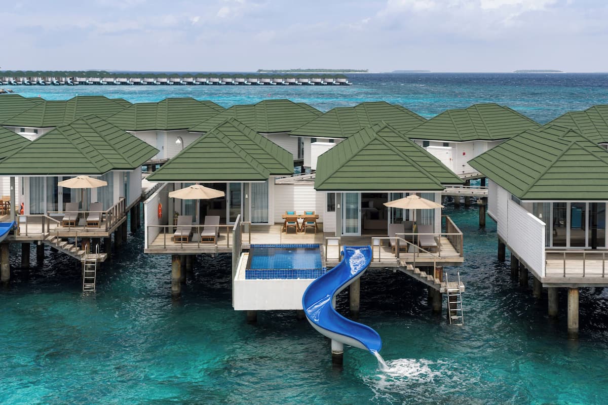 Overwater bungalows with a blue slide, clear turquoise water, and a distant island.