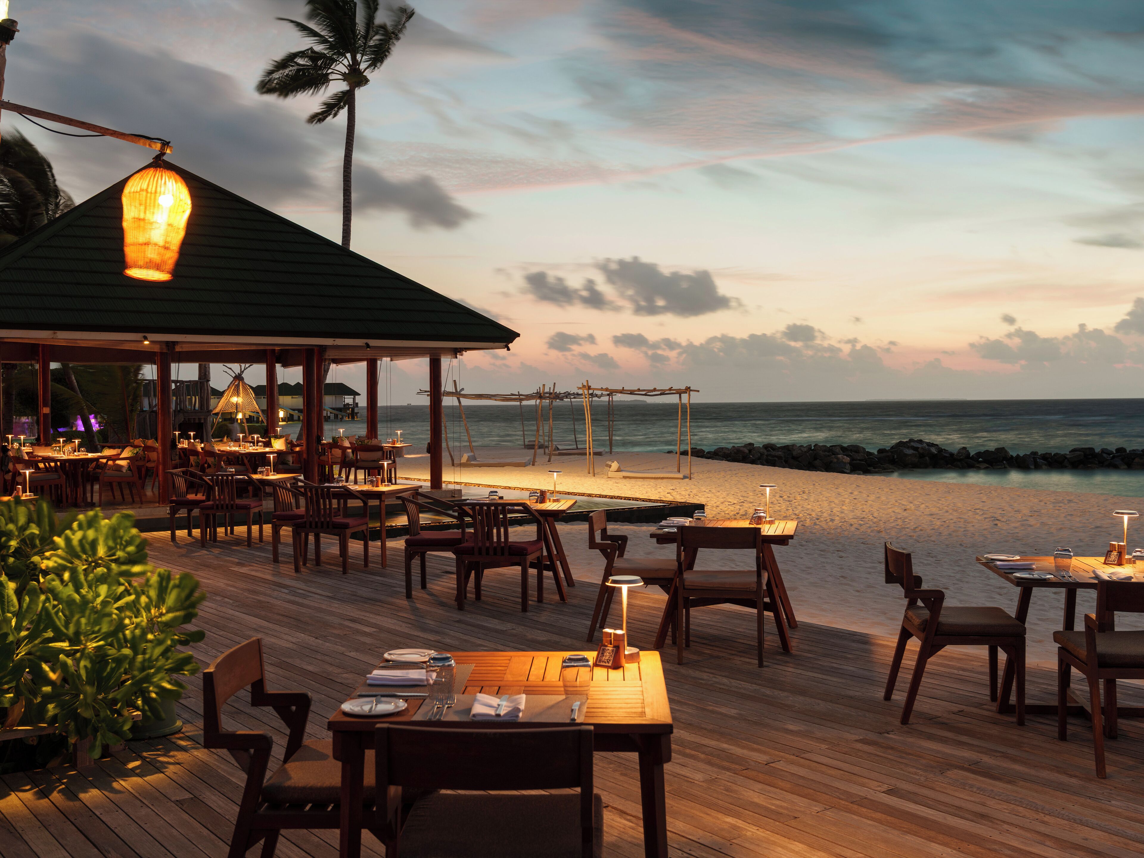 An outdoor dining area with wooden tables and chairs, overlooking a beach and ocean.