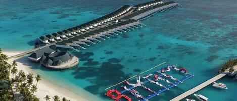 An overwater bungalows with a pier extending into turquoise waters, a beach with palm trees, and a water park area.