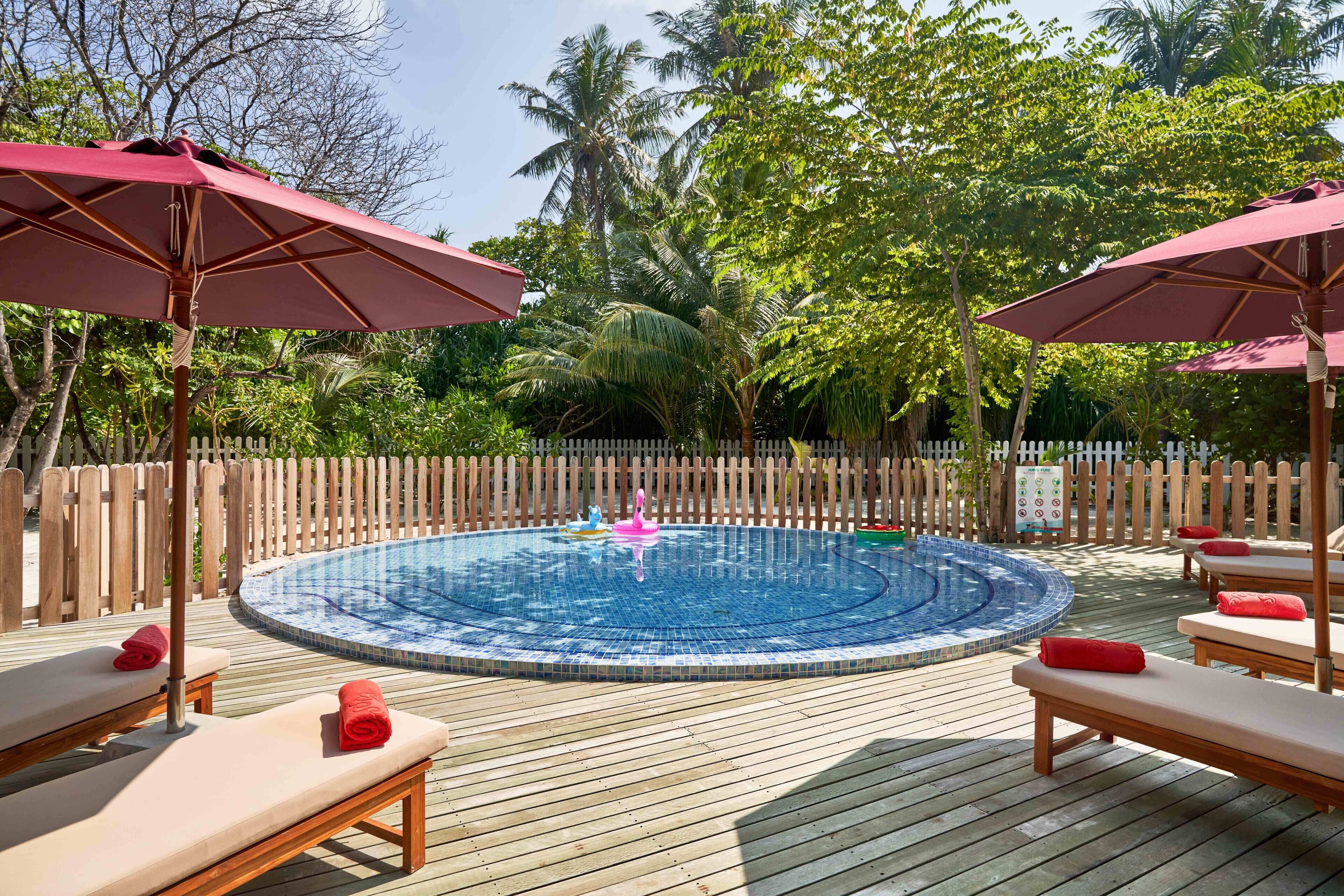 A circular hot tub surrounded by wooden deck lounge chairs with red towels, a wooden fence, and lush greenery in the background.