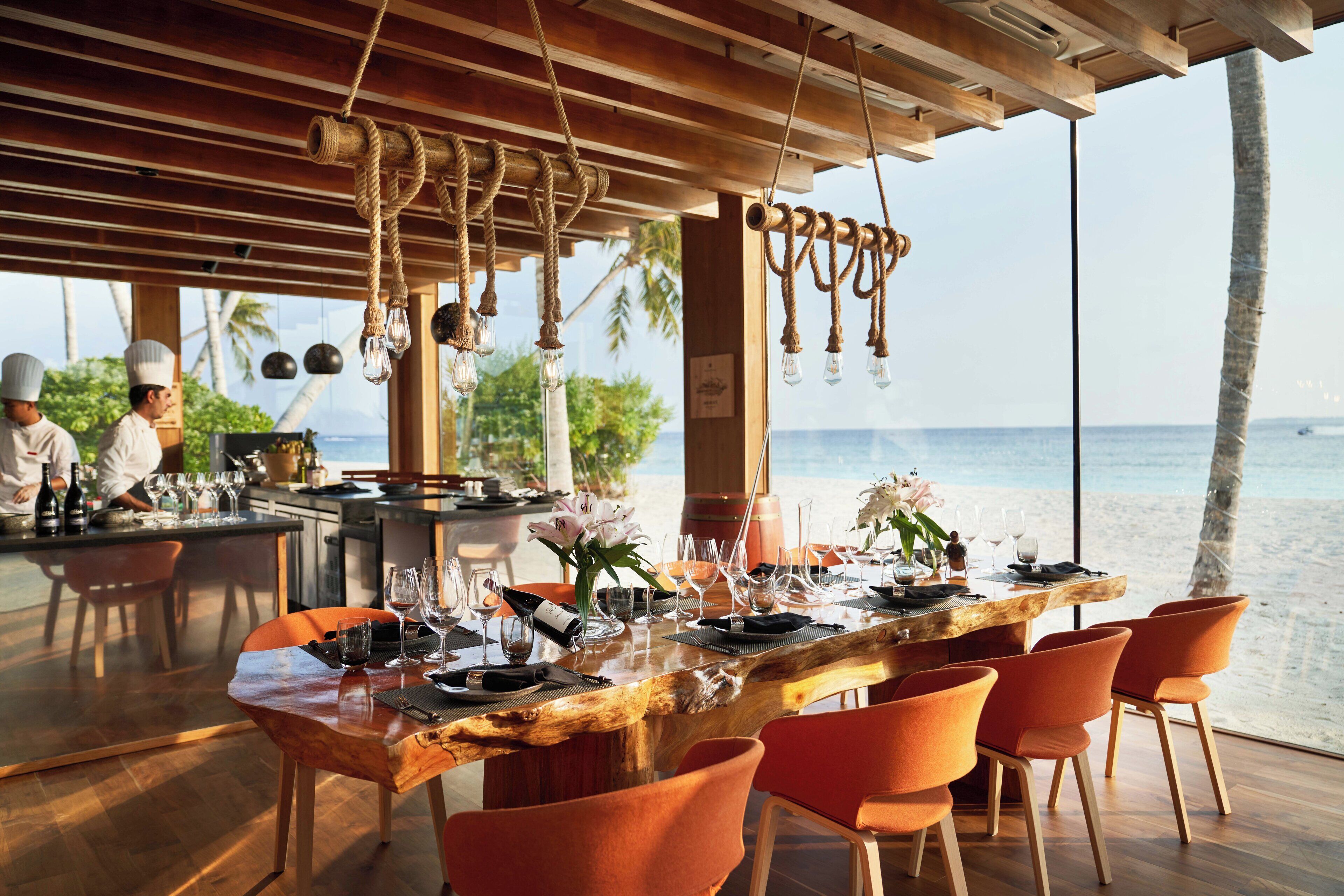 Un restaurante frente a la playa con mesa de madera, sillas anaranjadas y vista al océano.