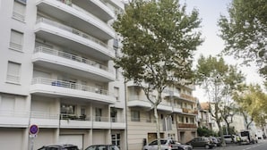 A multi-story residential building with balconies, parked cars, and trees along the street.