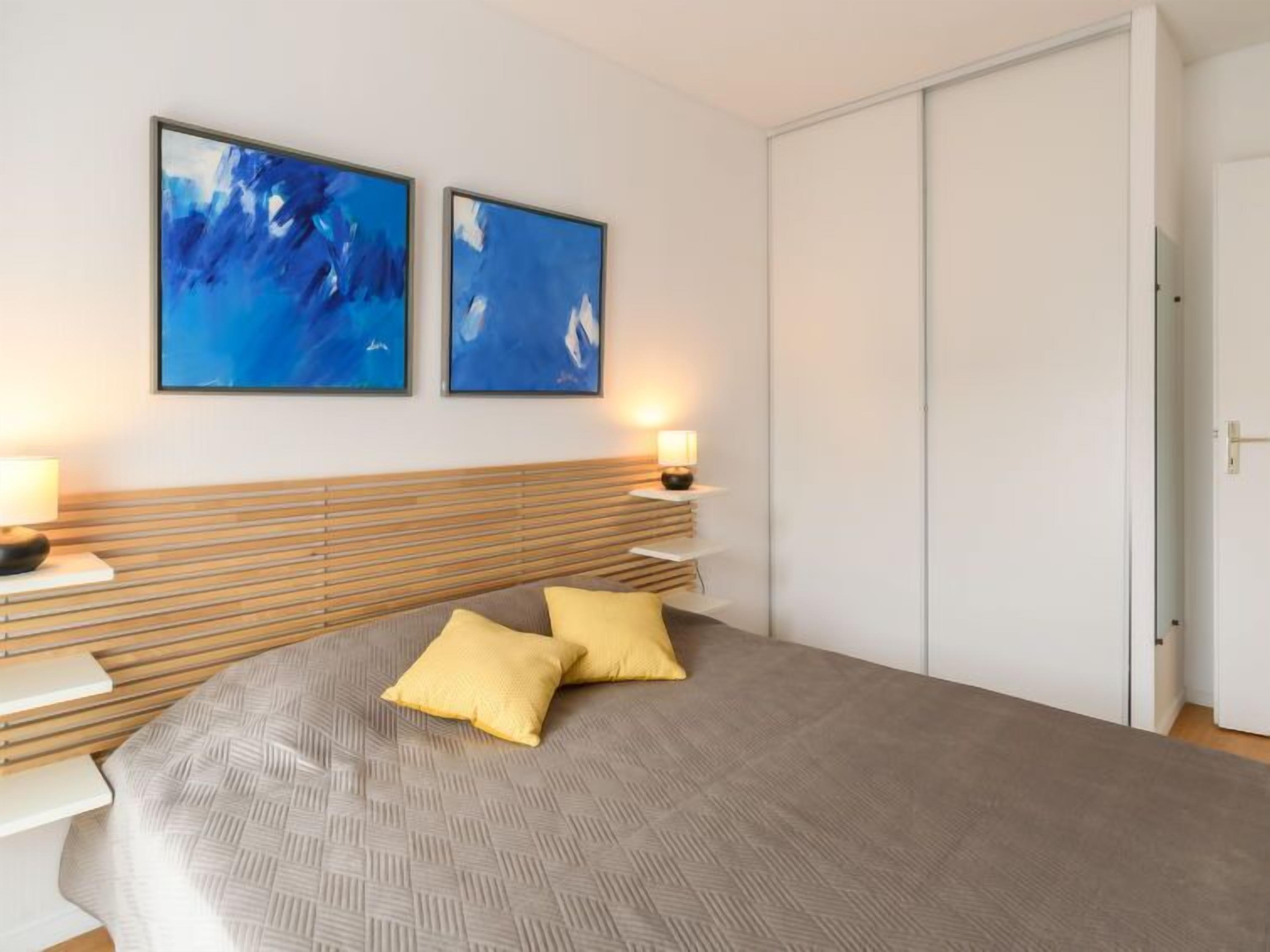 A bedroom with a bed, two wall-mounted paintings, bedside lamps, and a wardrobe.