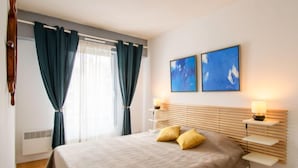 A bedroom with a bed, two bedside lamps, a window with curtains, and two wall-mounted paintings.