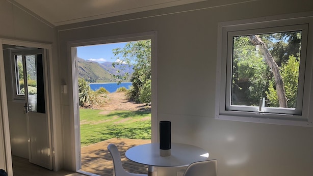 A room with a view of a lake and mountains, a table with chairs, and a door leading outside. - Lake Hawea Lakefront Cabin (Lake Hawea)