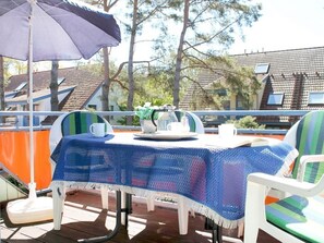 Eine Terrasse mit Tisch, bedeckt mit einer blauen Häkelttischdecke, weiße Stühle mit grün-blauen Streifenpolstern und einem Sonnenschirm.