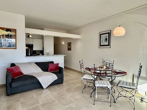 Uma sala de estar moderna com um sofá cinzento, uma mesa de jantar com quatro cadeiras e uma área de cozinha ao fundo.