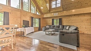 Ein geräumiges Wohnzimmer mit Holzwänden, einem grauen Sofa, einem Holztisch und einer Essecke mit Tisch und Stühlen.
