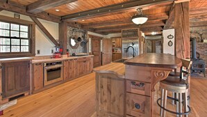 A rustic kitchen with wooden beams, a large island with stools, stainless steel appliances, and a brick wall. - Timeless Red Sox Retreat w/ Scenic Mountain Views! (Putney)