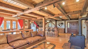 A cozy cabin interior with a wooden ceiling, a leather sofa, a glass coffee table, a kitchen area with a stove, and a stone fireplace. - Timeless Red Sox Retreat w/ Scenic Mountain Views! (Putney)