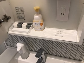 A bathroom with a white sink, a hand soap dispenser, a toothbrush holder with two toothbrushes, a bottle of Biore Marshmallow Whip, and a white towel on a shelf.