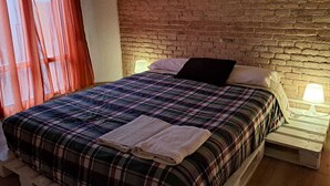 A bed with a plaid bedspread, two pillows, and a folded towel. There is a brick wall behind the bed and a window with curtains to the left. - Casa Antique Barcelona&Barca (Hospitalet de Llobregat)