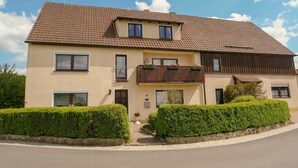 A two-story house with a tiled roof, a balcony with plants, and neatly trimmed hedges. - Ferienwohnung Liebenau - Ferienwohnung Wölfel (Gößweinstein)